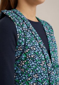 Prešívaný kvetinový vest s modrými, zelenými a bielymi kvetmi na námornickom pozadí. Má jemnú textúru a zaoblený design goliera.