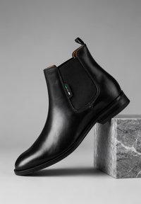 Crni Chelsea čizmić od kože, naslonjen na sivi mramorni blok, s elastičnim bočnim panelima i malom petljom na peti.