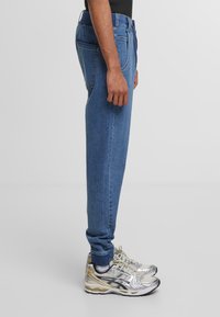 Blue denim jeans with a tapered fit, featuring a medium wash, five pockets, and contrasting stitching, paired with silver, gray, and gold sneakers.
