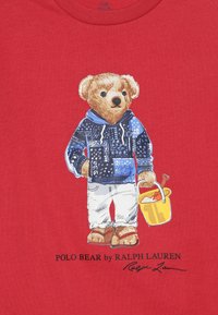Camiseta roja con un gráfico de un oso de peluche con una sudadera azul de patrón, pantalones blancos, sosteniendo un cubo amarillo con una pala.