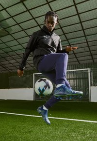 Athlete in black Puma jacket and navy pants, performing a kick with a black and white soccer ball on artificial turf. Blue cleats with green accents.