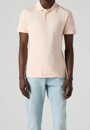 Mand iført lyserød kortærmet polotrøje og lyseblå jeans, stående mod en ensfarvet neutral baggrund.