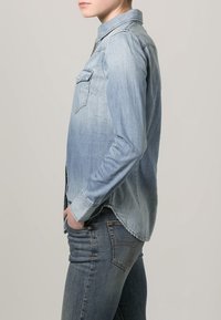 Chemise en denim bleu clair avec manches longues, col et poche poitrine avant. Le tissu présente une texture douce et un effet légèrement délavé.