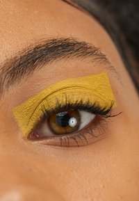 Close-up van een oog met gedurfde gele oogschaduw met een gestructureerde afwerking, omringd door donkere wimpers en een natuurlijke huidtint achtergrond.