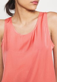 Korallfärgad ärmlös topp med en slät textur, rundad halsringning och lätt rynkad detalj vid kragen. Lättviktsmaterial.