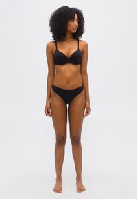 Femme aux cheveux bouclés portant un ensemble assorti de soutien-gorge et de culotte noirs, debout pieds nus devant un fond clair uni, regardant sur le côté.