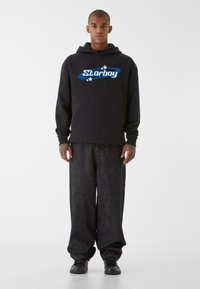 Felpa nera con cappuccio con logo blu "Starboy" e stelle bianche. Abbinata a pantaloni di jeans scuri a vestibilità ampia e sneakers nere.