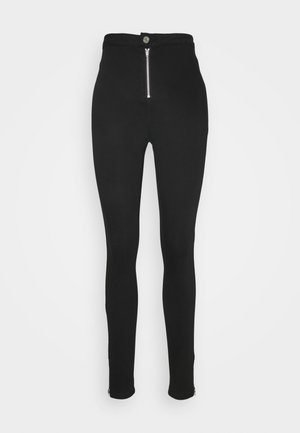 Pantalones negros de talle alto y ajuste slim, con cierre frontal de cremallera plateada y botón, y cremalleras en los bajos de los tobillos.