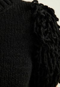 Maglione nero a maglia con un tessuto texturizzato e un accento frangiato ondulato su una spalla. Il materiale appare morbido e spesso.