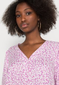 Blusa lilás com padrão floral branco, apresentando um decote em V e mangas soltas. Textura suave com tecido leve.