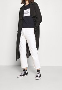 Persona che indossa un lungo cappotto nero, una felpa blu navy con la scritta "Tommy Hilfiger", pantaloni bianchi e sneakers alte nere Converse in piedi.