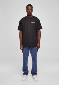 Svart bomull T-skjorte med løs passform, korte ermer, og en liten hvit logo på brystet, sammen med blå denimjeans og hvite sneakers.