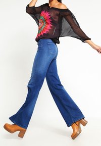 Czarna przezroczysta bluzka typu off-the-shoulder z kolorowym, okrągłym nadrukiem, zestawiona z rozszerzanymi niebieskimi dżinsami i brązowymi drewnianymi koturnami.