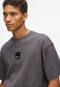 Pilka tekstūruota marškinė su apvaliu iškirpimu ir trumpomis rankovėmis, priekyje su mažą juodą etiketę.