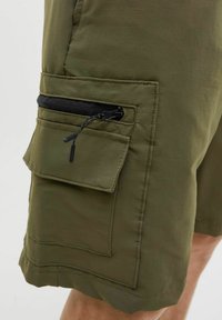 Shorts de culoare verde măslin cu un finisaj texturat, având o mare buzunar aplicat și un buzunar cu fermoar negru pe coapsă.