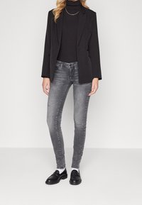 Blazer negro con un diseño ajustado, usado sobre un cuello de tortuga negro. Combinado con jeans ajustados grises y mocasines negros. Fondo liso.