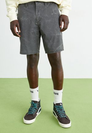 Shorts de denim gris con una textura ligeramente desgastada, combinados con zapatillas negras de caña alta con cordones turquesa y calcetines blancos hasta la altura del tobillo.