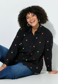 Lachende vrouw met krullend haar, grote oorbellen, een zwart shirt met kleurrijke sterren en een blauwe spijkerbroek, zittend tegen een witte achtergrond.