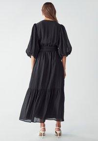 Black maxi dress with puffed sleeves, cinched waist, and tiered skirt. Made of textured fabric with a flowing design. White heeled sandals visible.