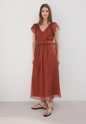 Vrouw draagt een roestkleurige maxi-jurk met korte mouwen en V-hals met geplooide taille, gecombineerd met zwarte platte sandalen met bandjes, staand tegen een witte muur.