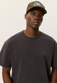 Σκούρο γκρι μπλουζάκι με στρογγυλή λαιμόκοψη και κοντά μανίκια. Διαθέτει καφέ λογότυπο "LES DEUX" στο στήθος. Χακί πράσινο καπέλο με λογότυπο.