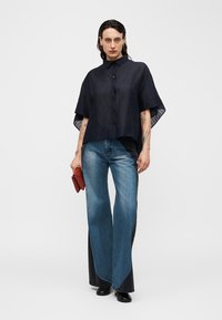 Žena s tmavými vlasy, která má na sobě volnou černou košili na knoflíky, džíny širokých nohavic v dvoubarevném provedení, černé boty a drží malou červenou kabelku.