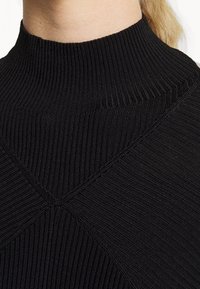 Svart ribbat topp med hög krage, tillverkad av ett strukturerat tyg, med en figurnära silhuett och distinkt sömdetaljering i ett chevronmönster.