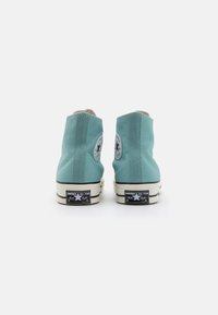Adidași înalți, dintr-un material textil de culoare teal deschis, cu un finisaj texturat. Talpa de cauciuc crem are o etichetă cu logo negru pe călcâi.