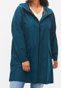 Teal vattentät jacka med dragkedja fram, justerbar huva och kontrasterande innerfoder, med sidofickor och tryckknappar i fållen.