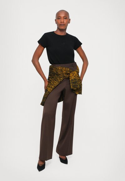 Black fitted t-shirt, brown trousers, and a patterned yellow-black scarf draped at the waist. Model wears black pointed heels.