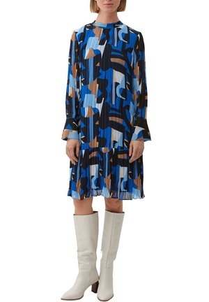 Femme portant une robe plissée à manches longues avec des motifs abstraits bleus, noirs, marron et blancs, associée à des bottes blanches jusqu'aux genoux.