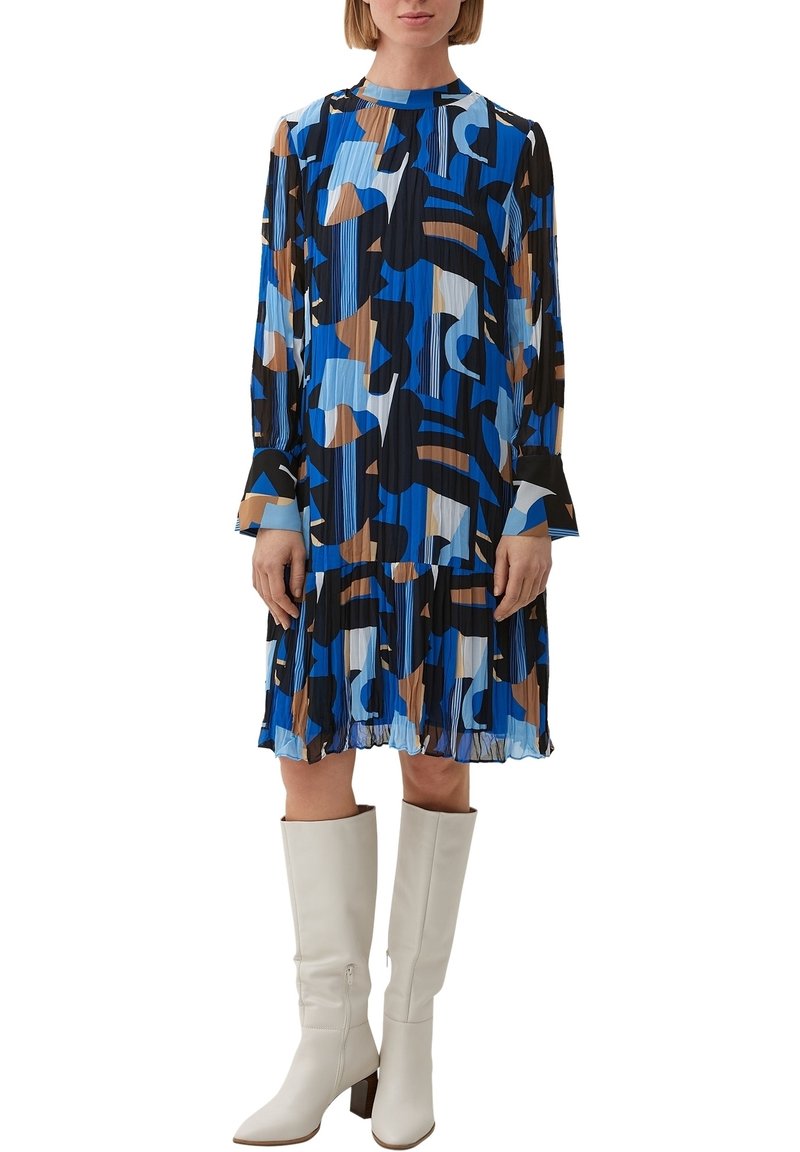 Femme portant une robe plissée à manches longues avec des motifs abstraits bleus, noirs, marron et blancs, associée à des bottes blanches jusqu'aux genoux.