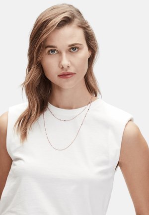 Gelaagde roségouden colliers met kralen en segmenten, gedragen over een witte mouwloze top. Glad textuur en delicaat ontwerp zijn opvallend.