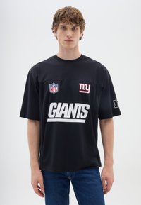 Maglietta nera a maniche corte con logo bianco "GIANTS", distintivo NFL e dettaglio "ny" rosso. Realizzata in tessuto morbido con una vestibilità comoda.
