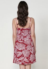 Rotes Kleid mit weißem Blumenmuster, ärmelloses Design und verstellbaren Trägern. Glatte Stoffqualität, leicht ausgestellter Schnitt, der bis zur Mitte des Oberschenkels reicht.