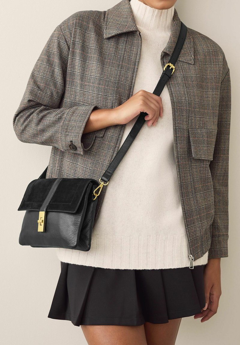 Schwarze Leder-Crossbody-Tasche mit einem Wildlederdeckel, ausgestattet mit einem goldenen Verschluss und einer glatten Oberfläche, kombiniert mit einer grauen karierten Jacke und einem cremefarbenen Pullover.