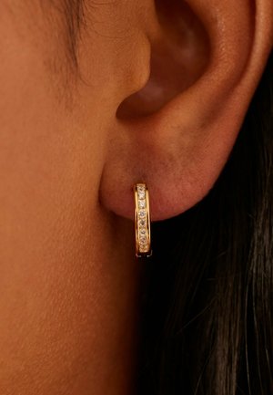 Boucle d'oreille créole en or avec des pierres précieuses claires incrustées portée sur une oreille à peau foncée, avec des cheveux noirs.