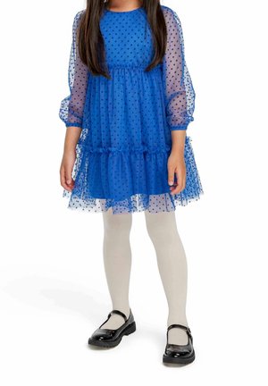 Niebieska sukienka z tiulu w groszki z bufkami przezroczystych rękawów i falbaną u dołu, zestawiona z kremowymi rajstopami i czarnymi lakierowanymi butami.