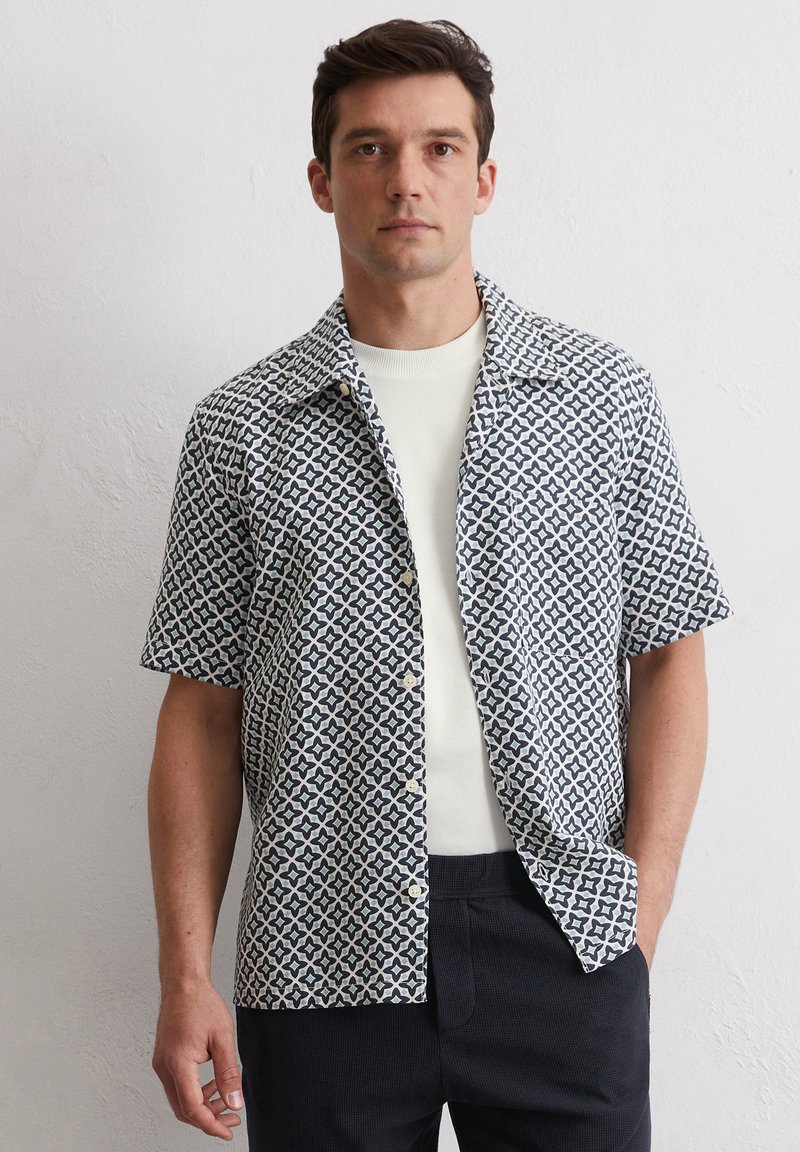 Camicia a maniche corte blu e bianca con motivo a stelle, chiusura con bottoni sul davanti e colletto; indossata sopra una maglietta a girocollo di colore chiaro.