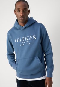 Felpa con cappuccio in misto cotone blu. Presenta un grande logo bianco, tasca a marsupio, polsini e orlo ribattuti. La texture appare liscia.