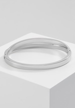 Bracelet jonc en argent lisse et poli avec un fermoir à charnière, posé sur un piédestal géométrique blanc sur un fond gris clair.