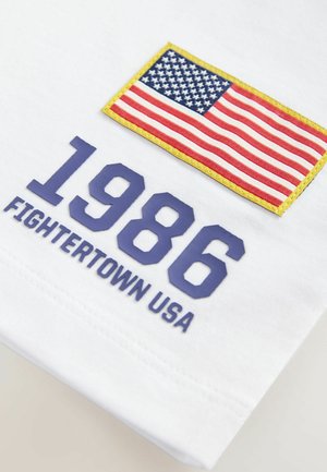 T-shirt en coton blanc avec un patch de drapeau américain, le texte bleu "1986" et "FIGHTERTOWN USA" en bleu, entouré d'une bordure jaune sur le drapeau.