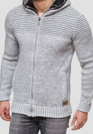 Mand iført lysegrå strikket trøje med lynlås, ribstrikkede skuldre, ærmegab, kant og sidelommer over grå jeans.