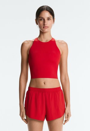Mladá žena s tmavými vlasmi má na sebe červený bezrukávový crop top a voľné červené šortky, stojí čelom dopredu na svetlom jednofarebnom pozadí.
