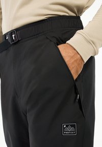 Zwarte outdoor broek van zacht stof, met een elastische tailleband, een ritszak en een klein logo-opdruk op de dij.