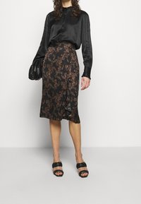 Blouse en satin noir associée à une jupe noire ornée d'un motif abstrait marron, avec des détails froncés sur le côté, et des sandales noires à lanières.