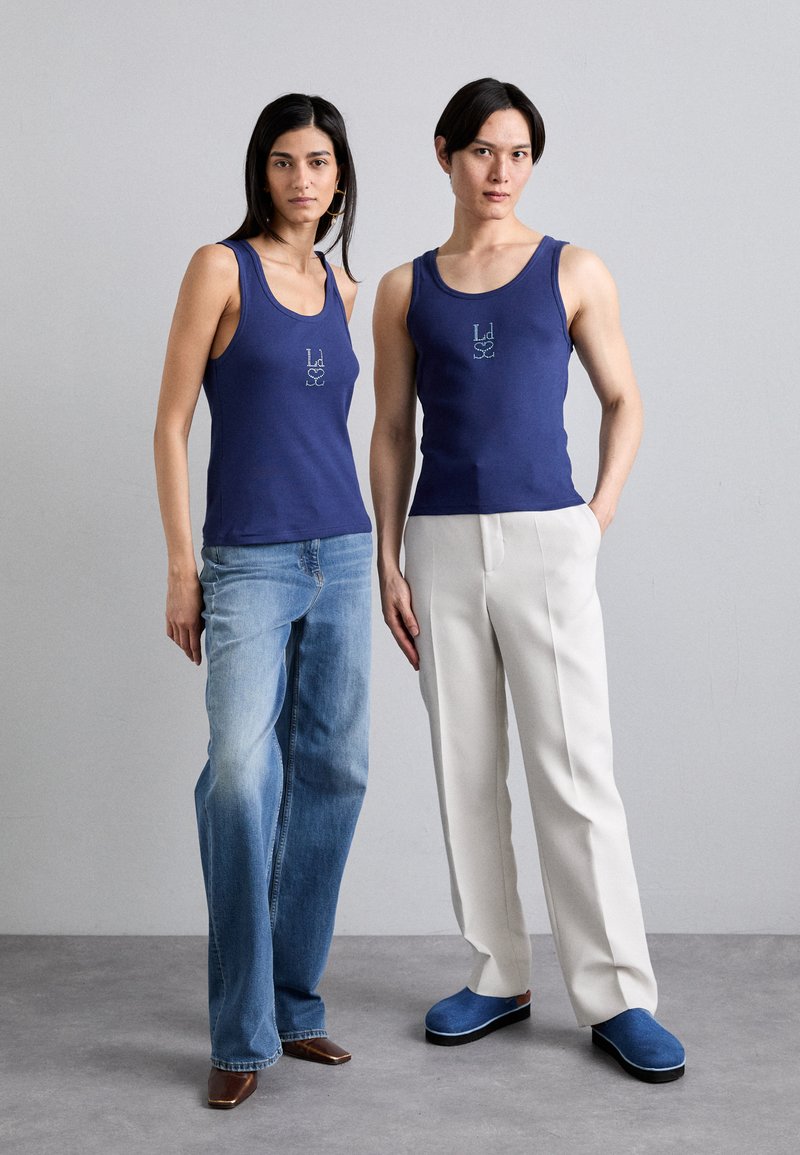 Blaue Baumwolltanktops mit einem kleinen Logo auf der Brust, kombiniert mit hellen Jeans und weißen Hosen. Die Models tragen moderne Schuhe in Blau und Braun.