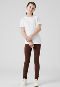 Witte t-shirt met korte mouwen gecombineerd met bruine leggings, beide gemaakt van een gladde stof. Het model draagt witte sneakers.