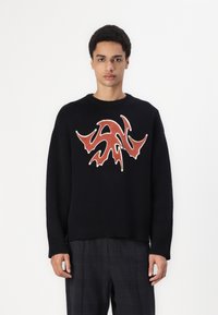Sort sweater med lange ærmer, ribbet søm og manchetter. Har et fremtrædende rødt abstrakt design på forsiden. Bæres med ternede bukser.