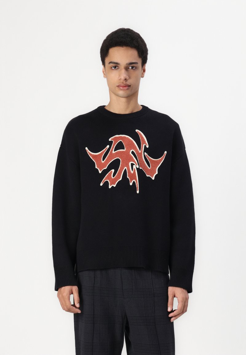 Sort sweater med lange ærmer, ribbet søm og manchetter. Har et fremtrædende rødt abstrakt design på forsiden. Bæres med ternede bukser.